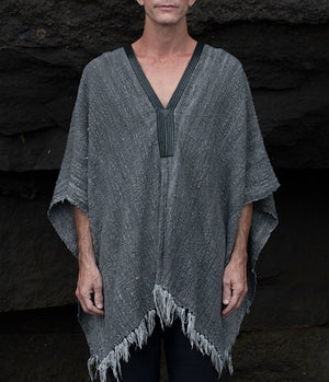 Person wearing a gray poncho with fringe details against a dark stone wall.