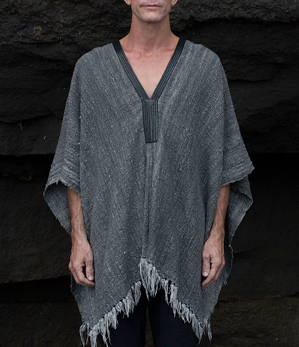 Person wearing a gray poncho with fringe details against a dark stone wall.