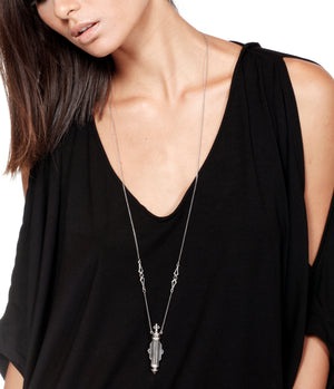 Person wearing a black top with a long silver necklace featuring a art nouveau perfume bottle pendant.
